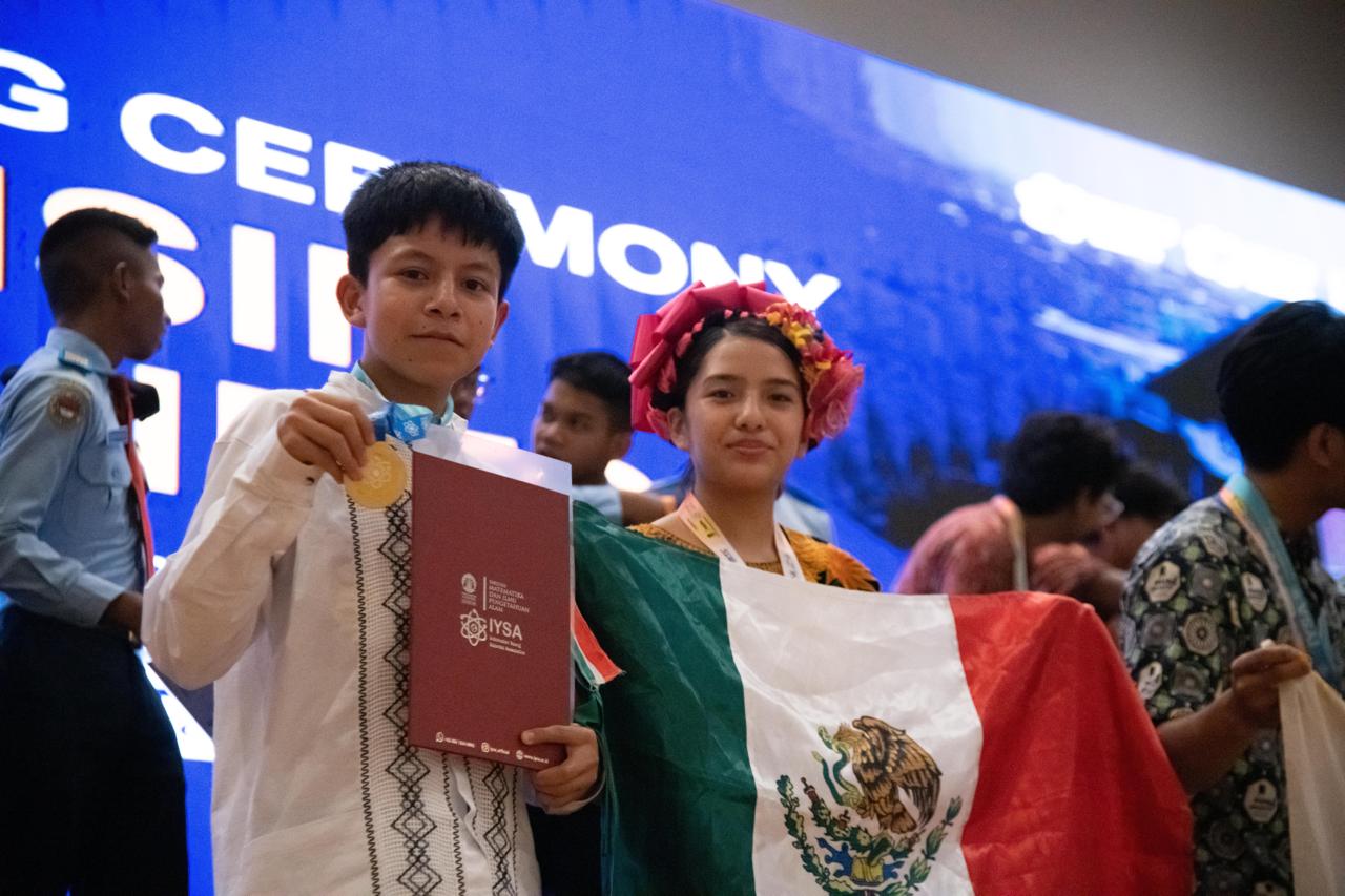 Brayan muestra su medalla y su carpeta de IYSA junto a Vioreny, que sostiene la bandera de México, durante la ceremonia de clausura