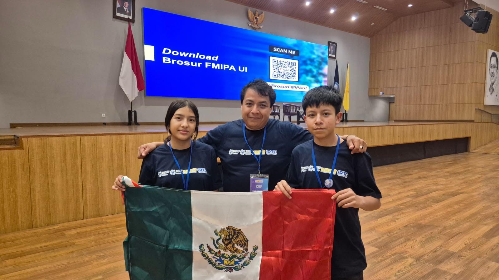 Vioreny, el profesor Francisco Javier Cruz Hernández y Brayan sostienen la bandera de México en un auditorio
